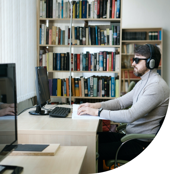 Um homem branco está sentado em uma cadeira, de frente para um computador. Ele usa óculos escuros e faz uma leitura em braile. Também usa headphones. Ao fundo, uma estante de livros.